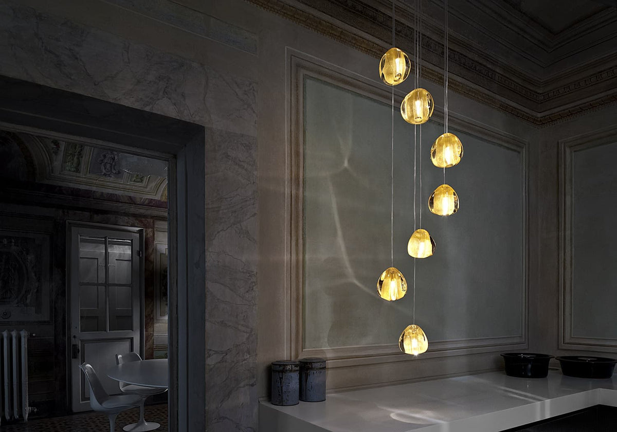 Warm yellow glass heads of this Clérachic pendant hang down a panelled wall, with a white counter and a doorway at the left.