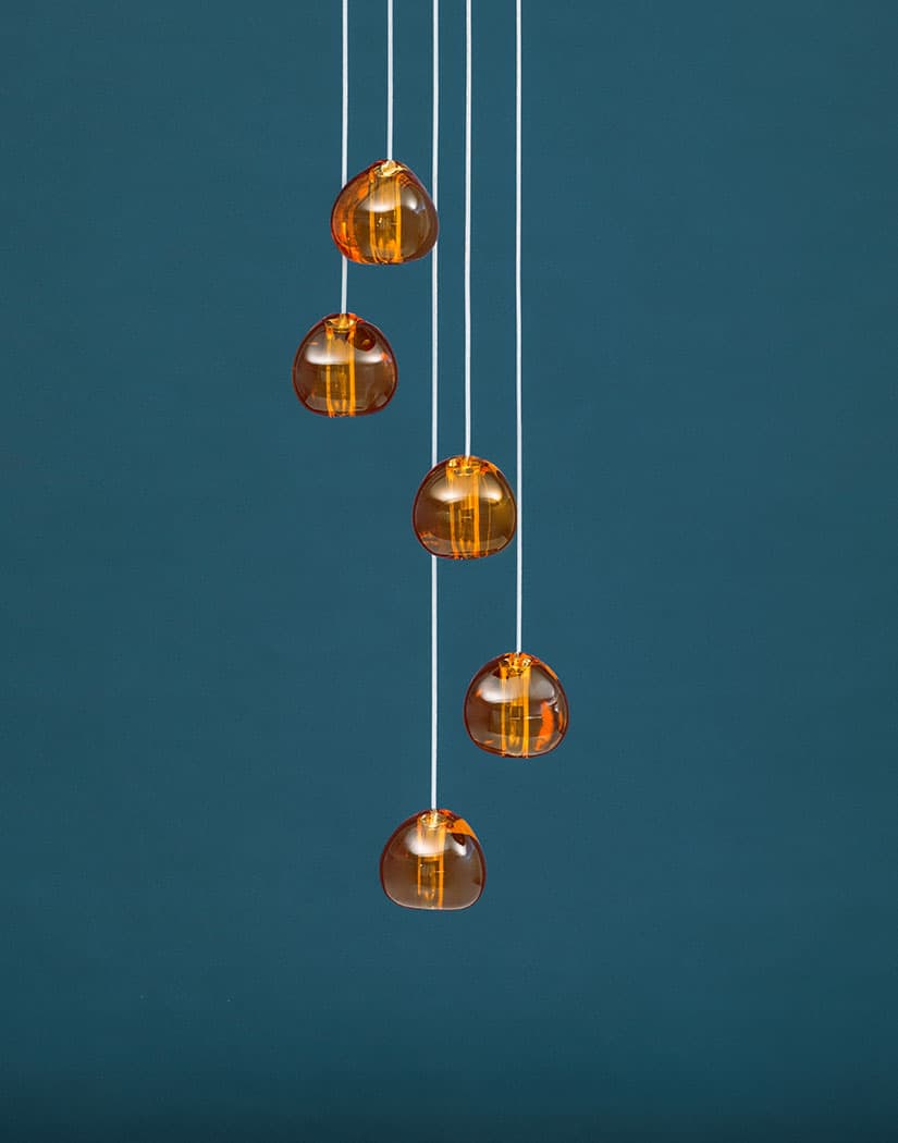 Five amber glass heads on this Clérachic pendant hang down a plain teal wall, each one glowing with a warm light.