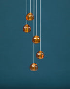 Five amber glass heads on this Clérachic pendant hang down a plain teal wall, each one glowing with a warm light.