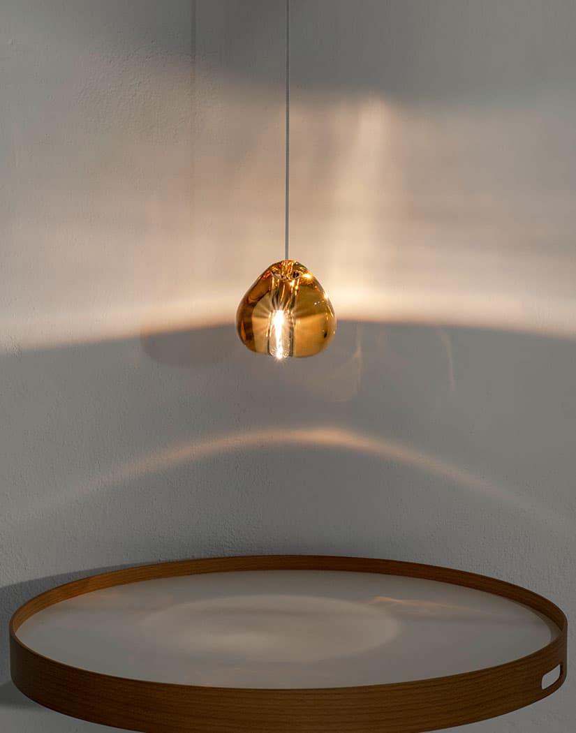 A single amber glass head from this Clérachic pendant glows above a round wooden tray, with soft light curving across the wall.