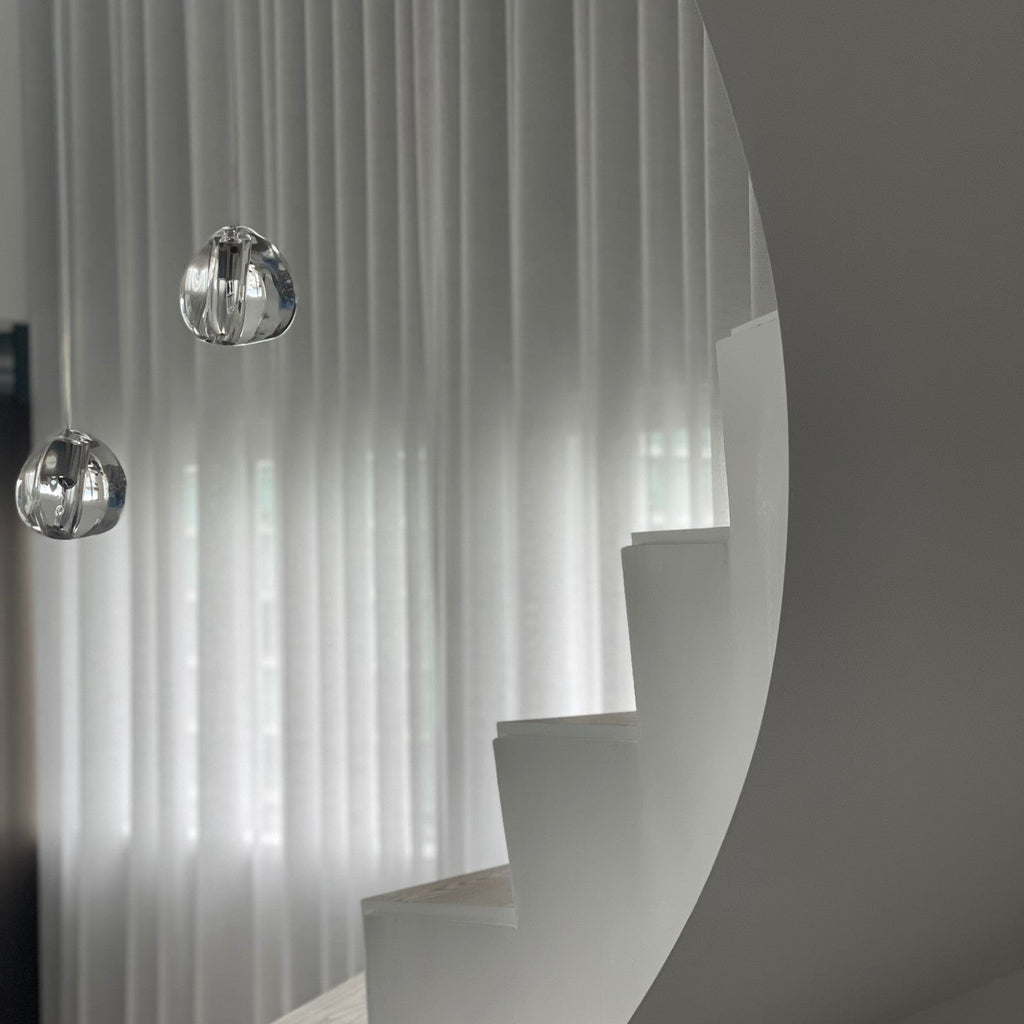 Two clear glass heads from this Clérachic pendant hang in front of sheer curtains, beside a curved white staircase.
