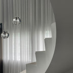 Two clear glass heads from this Clérachic pendant hang in front of sheer curtains, beside a curved white staircase.