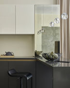 Clear glass heads hang near the top of this kitchen scene, over a dark counter with a teapot, a bowl and a black stool, from a Clérachic pendant.