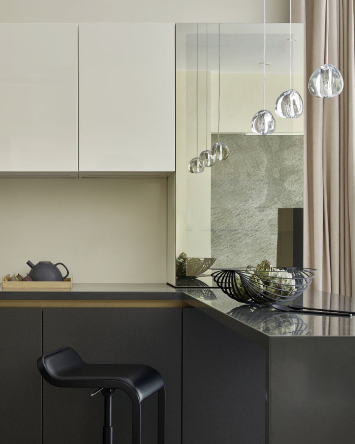Clear glass heads hang near the top of this kitchen scene, over a dark counter with a teapot, a bowl and a black stool, from a Clérachic pendant.