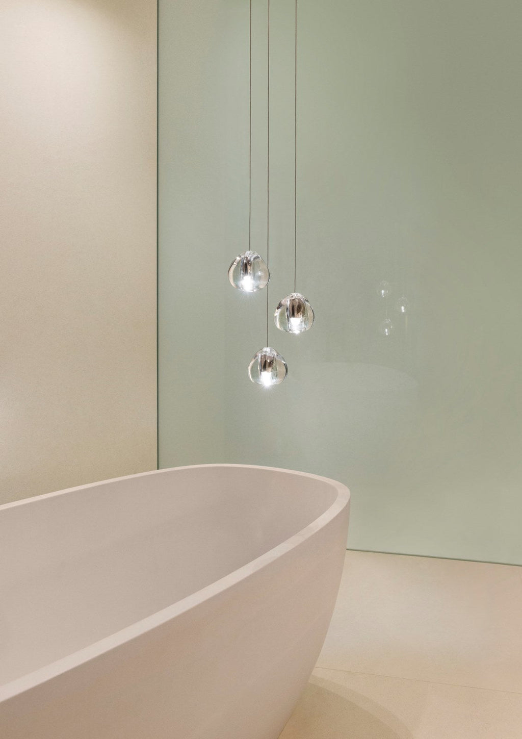 A smaller group of clear glass heads on this Clérachic pendant hangs close to a pale bath and a green-grey wall.