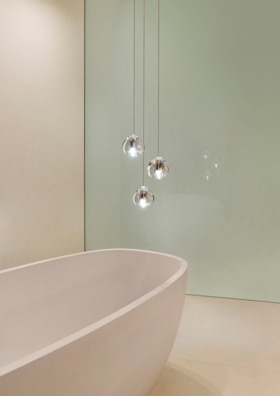 A smaller group of clear glass heads on this Clérachic pendant hangs close to a pale bath and a green-grey wall.