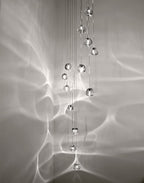 Clear glass heads of this Clérachic pendant hang in a tall column against a pale grey wall, with a web of light ripples around them.