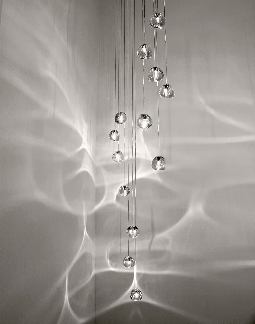 Clear glass heads of this Clérachic pendant hang in a tall column against a pale grey wall, with a web of light ripples around them.