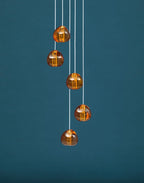 Five amber glass heads on this Clérachic pendant glow in a staggered line against a flat teal background.