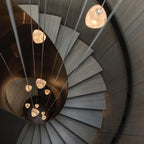 Looking down a spiral staircase, this Clérachic pendant's glowing glass heads are seen against a dark copper-toned stairwell.
