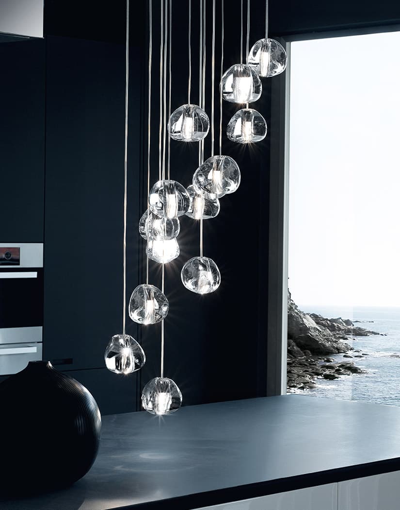 A tall view of the clear glass heads of this Clérachic pendant hanging in a dark kitchen with a window onto the sea.