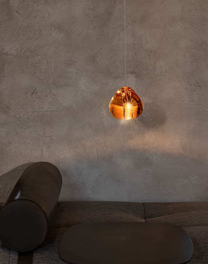 One amber glass head from this Clérachic pendant glows against a rough concrete wall above a low dark sofa.