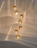 Seven amber glass heads of this Clérachic pendant glow on a warm cream wall that is marked with soft light ripples.