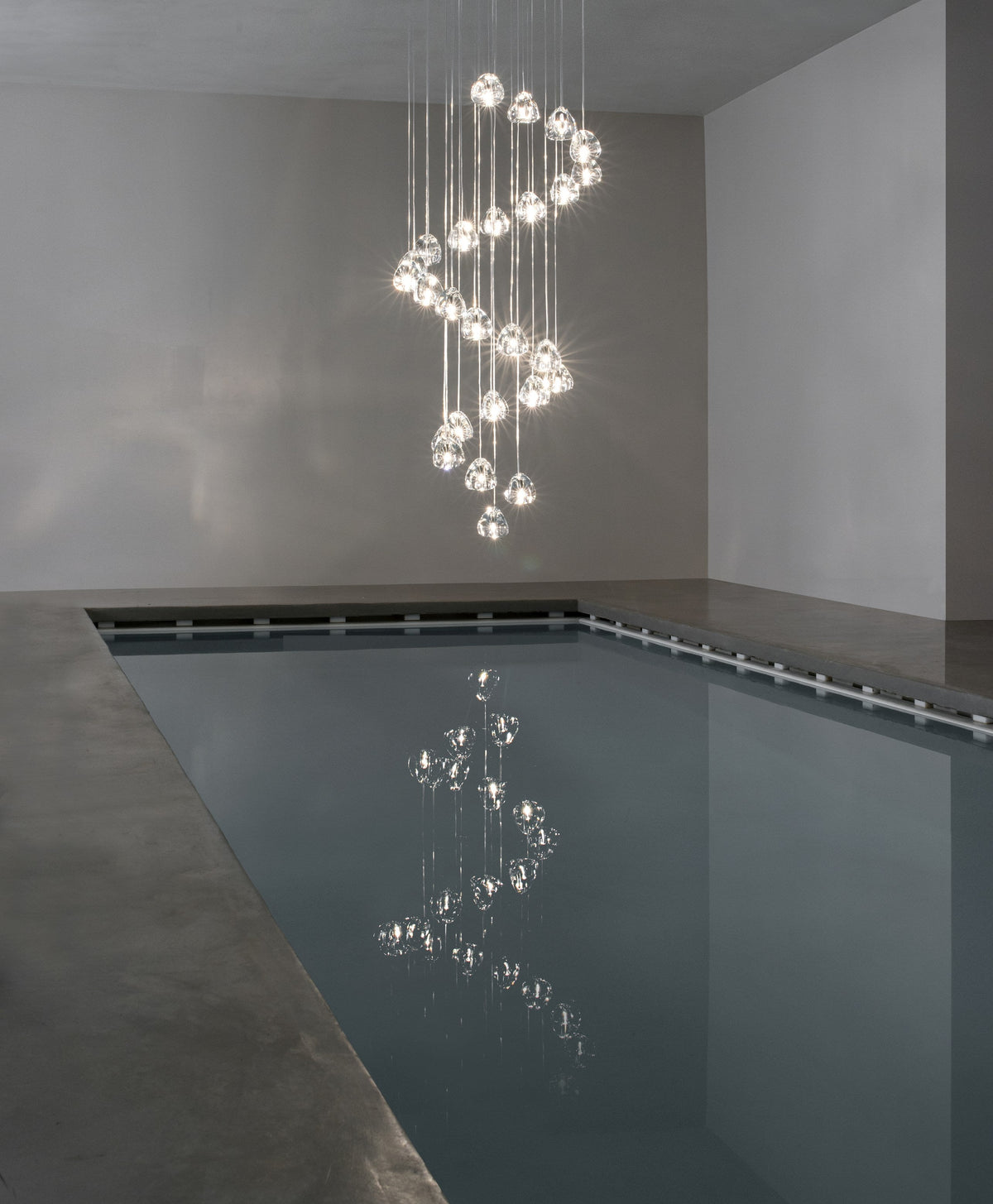 A spiral column of clear glass heads on this Clérachic pendant hangs over a long, still indoor pool in a plain concrete room.