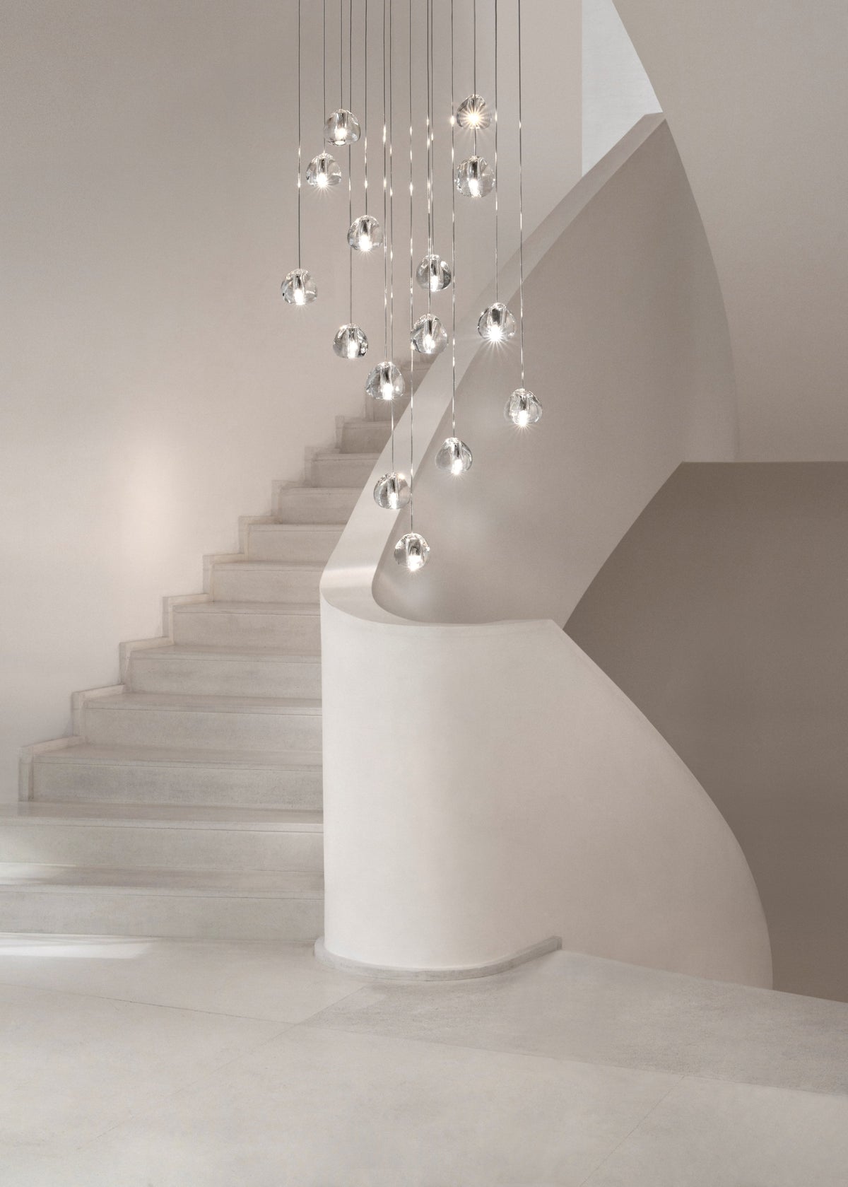 This Clérachic pendant hangs as a cluster of clear glass heads, high above a white staircase with a curved wall.