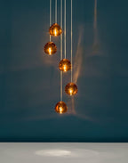 Five amber glass heads on this Clérachic pendant glow against a dark teal wall, with a pale floor reflecting light below.