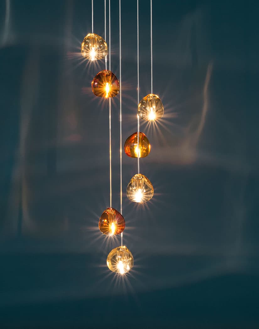 Amber and pale gold glass heads of this Clérachic pendant hang in a line, each with a bright starburst of light, against deep teal.