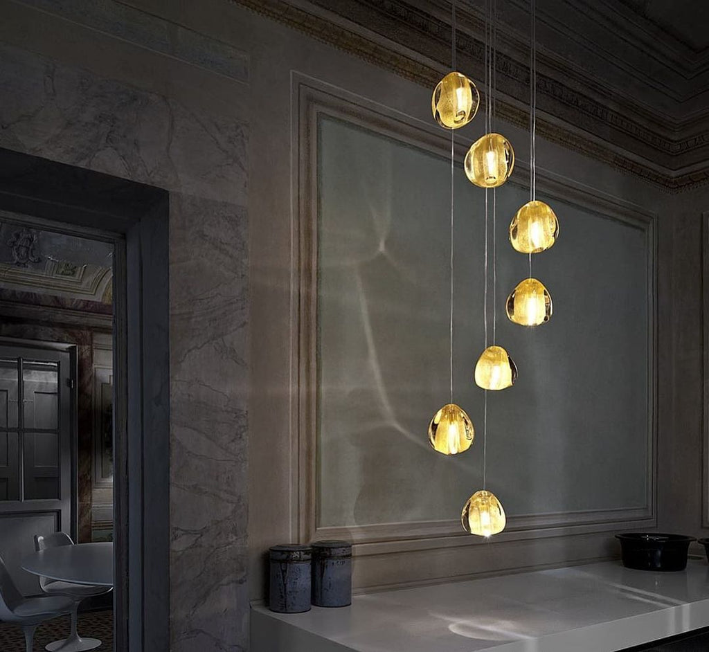 Seven warm yellow glass heads of this Clérachic pendant hang in front of a moulded wall panel in a dim, elegant room.