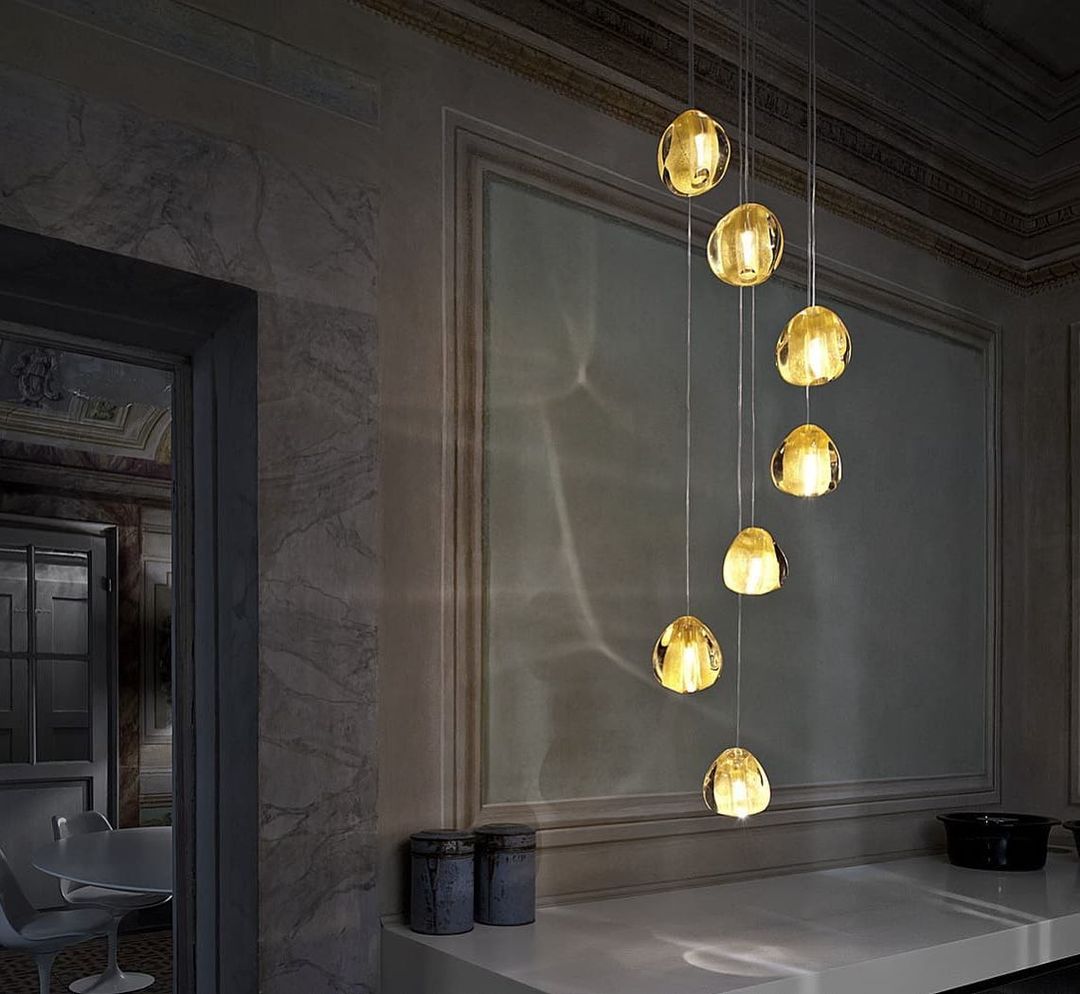 Seven warm yellow glass heads of this Clérachic pendant hang in front of a moulded wall panel in a dim, elegant room.
