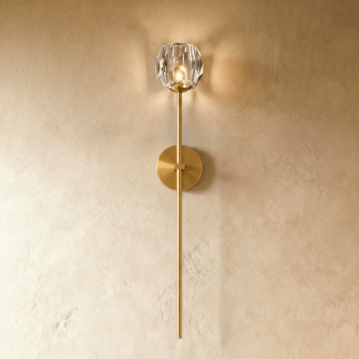 Clérachic gold wall sconce, a faceted crystal shade lit warm on a long brass-toned stem and round wall plate.