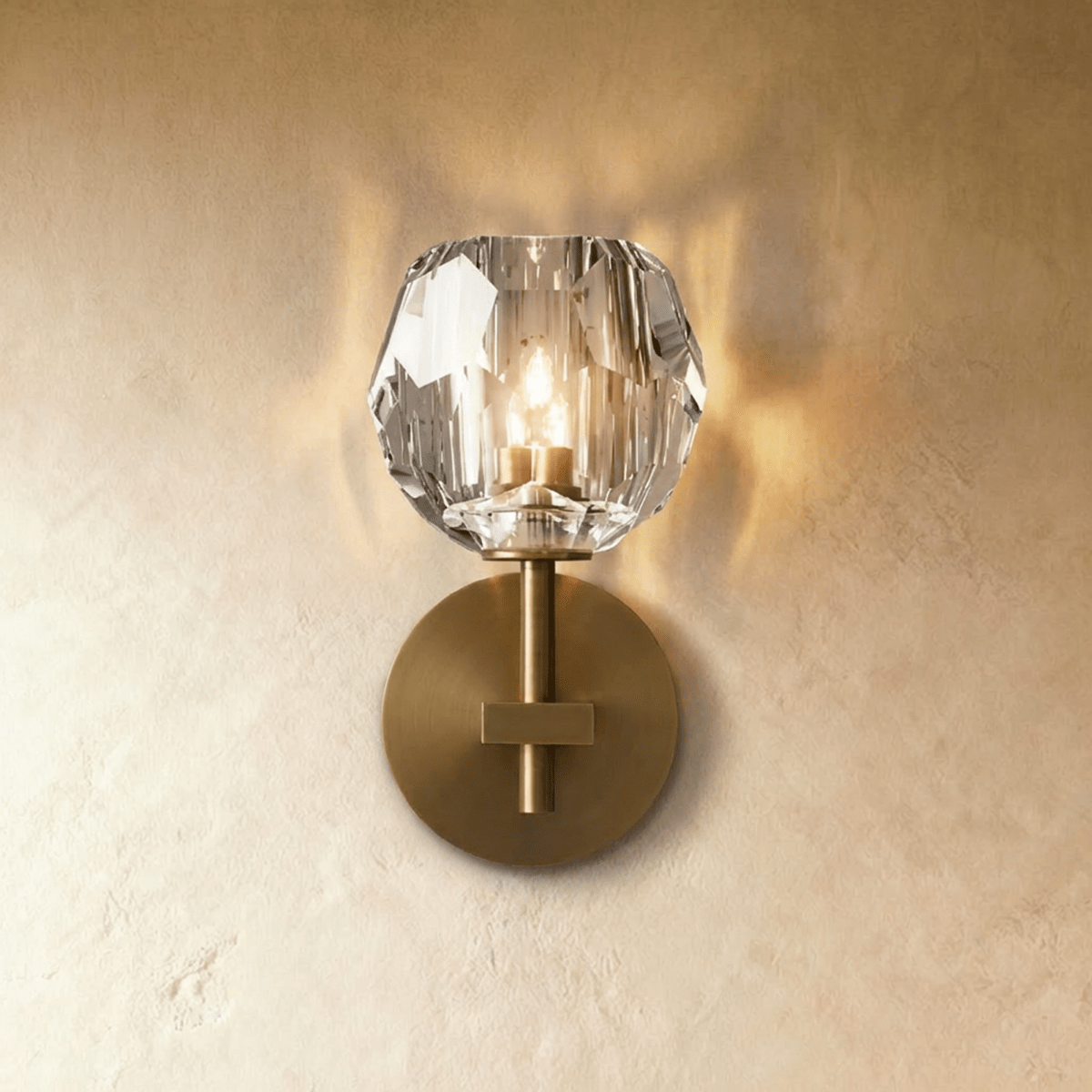 Clérachic short gold wall sconce, a faceted crystal shade glowing warm above a brushed brass round wall plate.