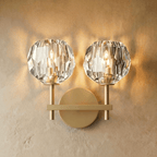 Clérachic two-head gold wall sconce, twin faceted crystal shades glowing on a brass crossbar and round wall plate.