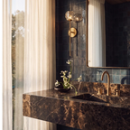 Clérachic gold wall sconce with a crystal shade on a dark glazed tiled wall beside a sheer curtain and framed mirror.