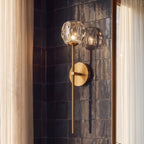 Clérachic gold wall sconce, close view of its crystal shade and brass stem against dark glazed tiles and a sheer curtain.