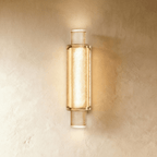 Clérachic slim vertical wall light of textured crystal glass in a gold frame, glowing warm against a plastered wall.
