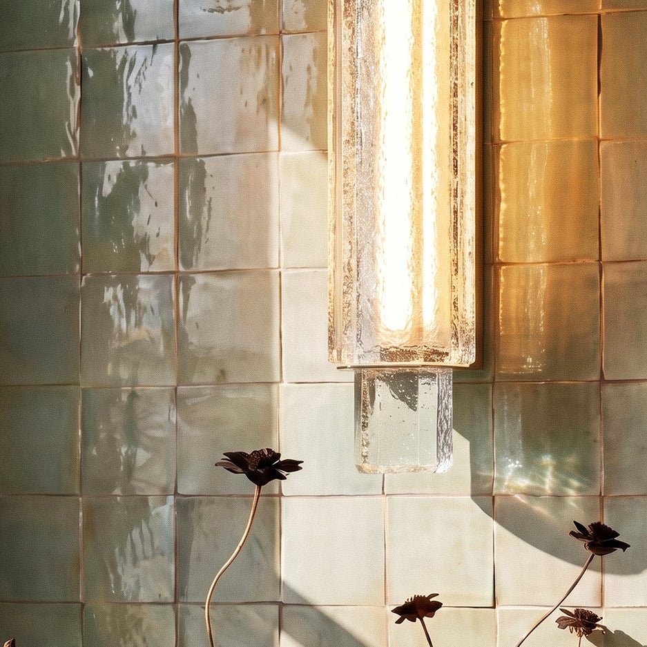 Close view of a Clérachic textured crystal wall light glowing warm against glossy green tiles, with dark flower stems below.