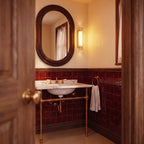 Clérachic slim gold and crystal wall light lit beside an oval mirror in a tiled room with deep red tiles and brass taps.