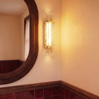 Clérachic gold-framed crystal wall light glowing warm on a cream wall beside an oval wooden mirror and red tile.