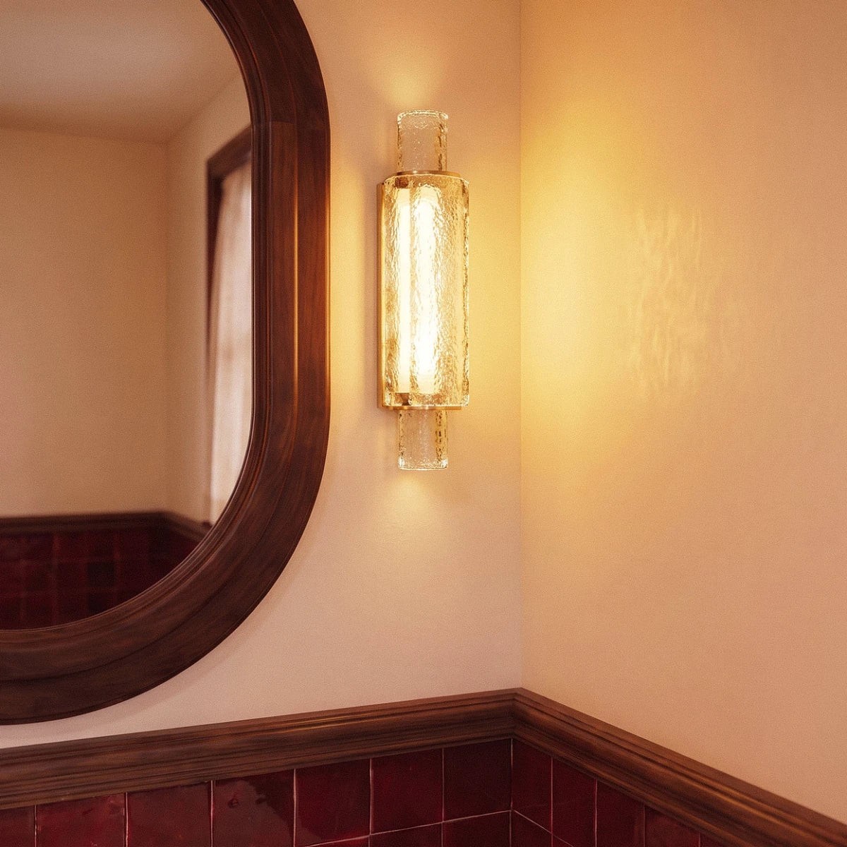 Clérachic gold-framed crystal wall light glowing warm on a cream wall beside an oval wooden mirror and red tile.