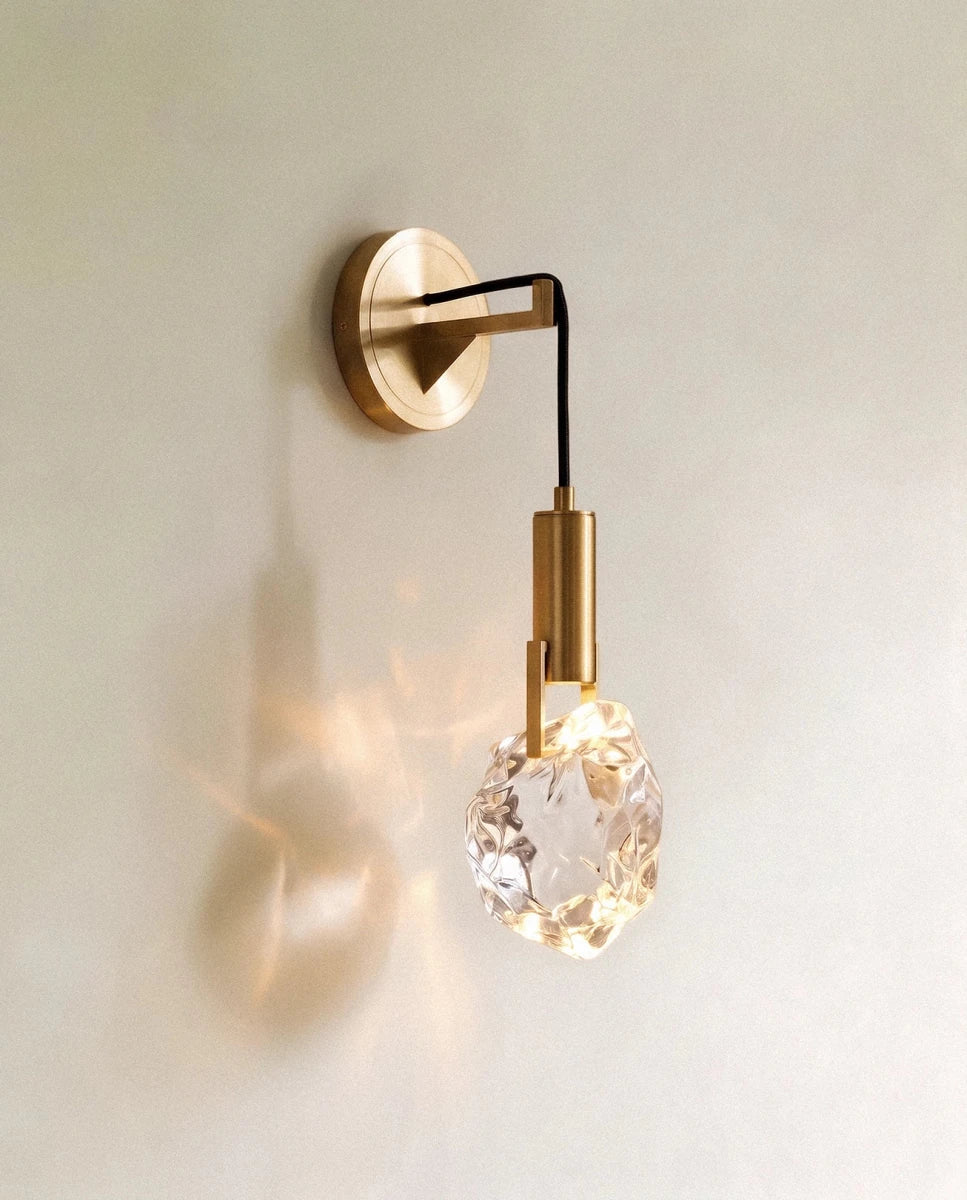 Clérachic crystal wall sconce, a rough-cut glass stone glowing warm on a slim brass arm against a plain wall.