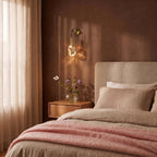 Clérachic crystal wall sconce glowing warm beside a bed, its brass arm catching evening light on a plaster wall.