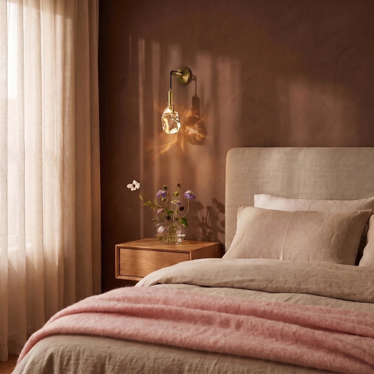 Clérachic crystal wall sconce glowing warm beside a bed, its brass arm catching evening light on a plaster wall.