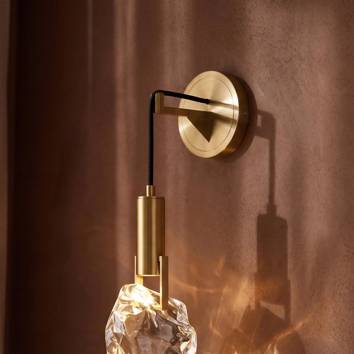 Close-up of the Clérachic crystal wall sconce's brass mount and glass stone against a warm curtained backdrop.