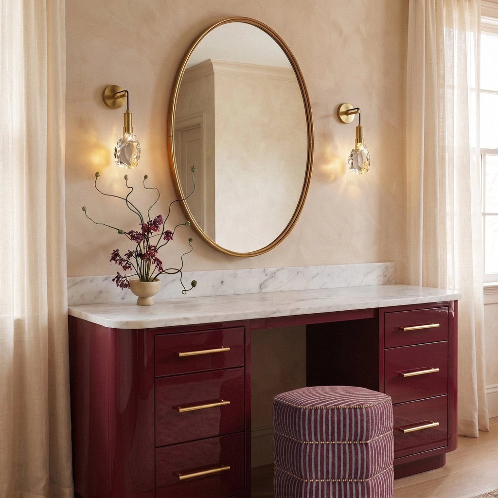 Two Clérachic crystal wall sconces flank an oval mirror above a glossy burgundy vanity with a marble top.
