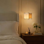 Clérachic cube pendant glowing over a bedside table with a candle, a reed diffuser, books and white peonies.