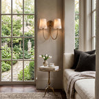 Two-light Clérachic alabaster wall light glowing beside a tall garden window, with a small brass side table and cream sofa below.