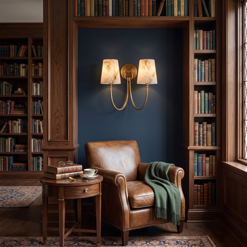 Two-light Clérachic wall sconce lighting a blue reading nook between bookshelves, with a tan leather armchair and a green throw.