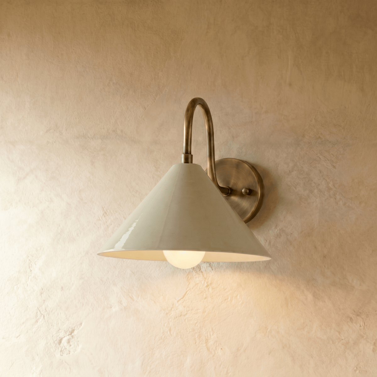 Clérachic cone-shade wall sconce in vintage brass with a gooseneck arm, mounted on a plaster wall in studio light.