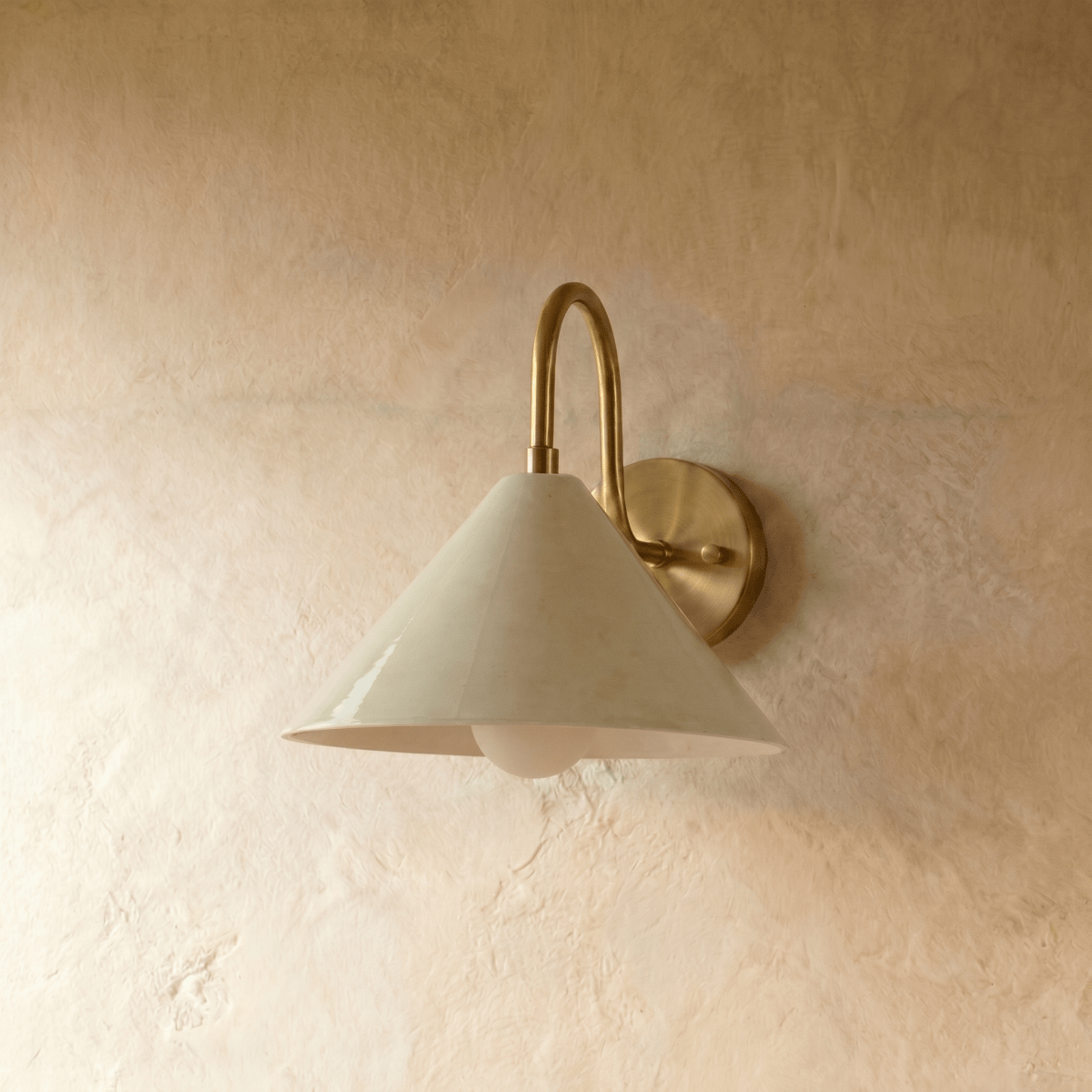 Clérachic cone-shade wall sconce in polished brass with a gooseneck arm, mounted on a plaster wall in studio light.