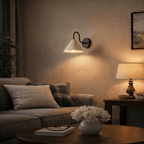 Clérachic matte black cone-shade wall sconce lit above a sofa with cushions, a framed print and a table lamp nearby.