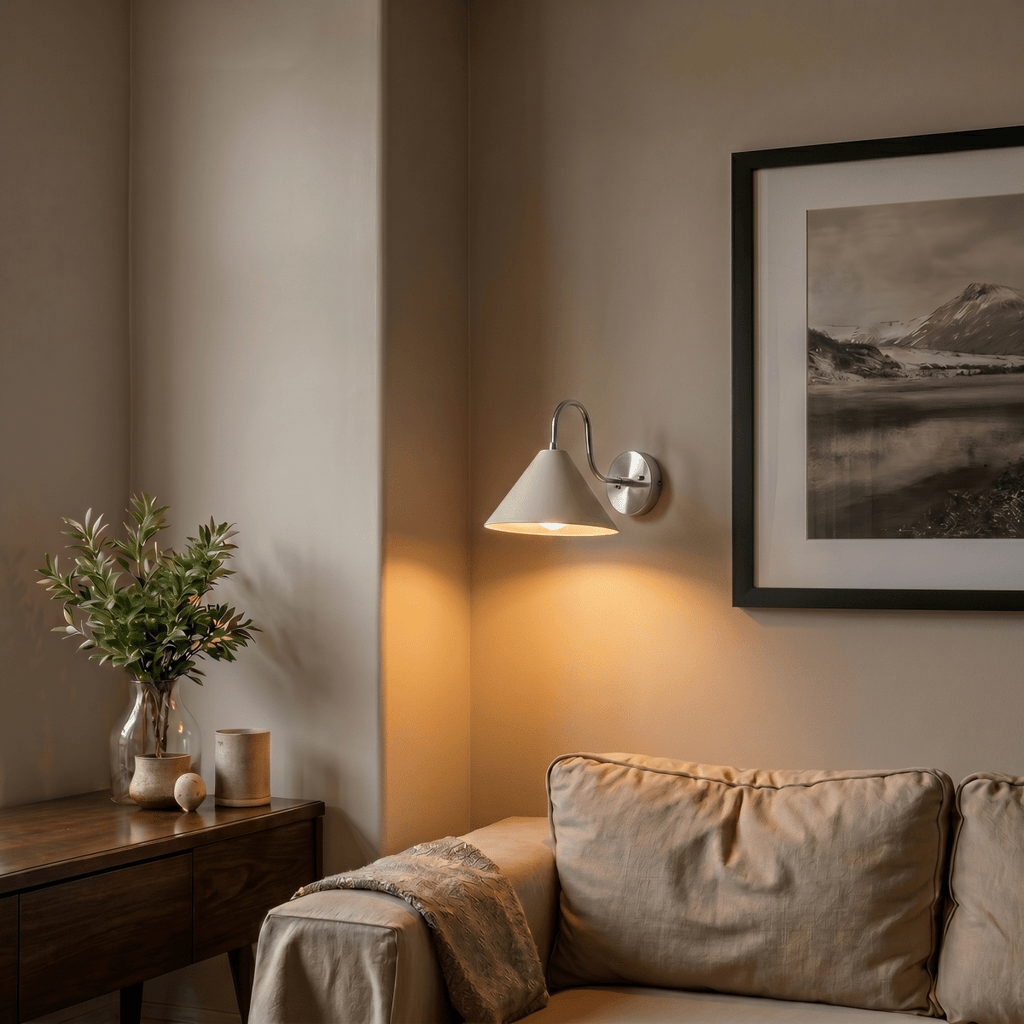 Clérachic chrome cone-shade wall sconce lit in a living room corner beside a framed landscape print and a sofa.