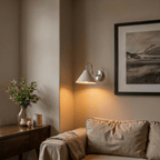Clérachic chrome cone-shade wall sconce lit in a living room corner beside a framed landscape print and a sofa.