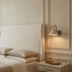 Clérachic vintage brass cone-shade wall sconce lit above a panelled bedroom wall beside a marble-top nightstand.