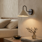 Clérachic matte black cone-shade wall sconce lit above a bedside table with a bowl, a vase and stacked books.