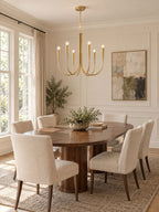 A Clérachic chandelier with brass-toned arms above an oval wooden dining table and cream chairs, beside a tall window with a drawn curtain.