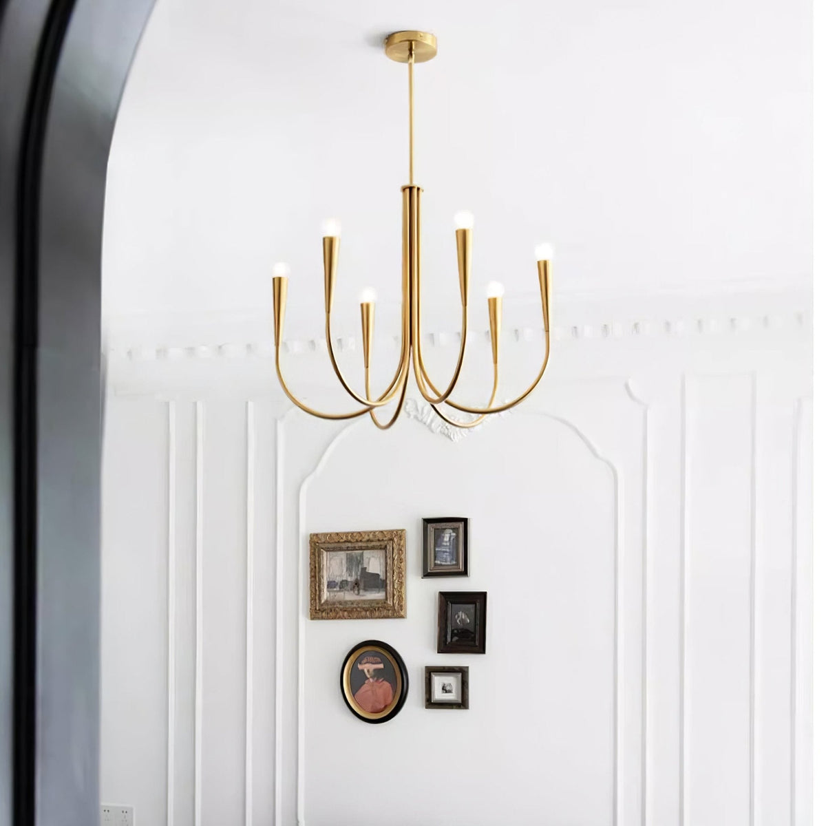 A smaller Clérachic chandelier with brass-toned arms against a white panelled wall, with a cluster of framed pictures below it.